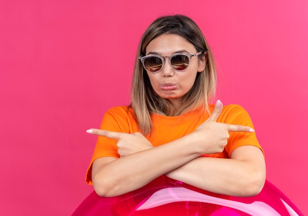 Foto gratuita una giovane donna triste in una maglietta arancione che indossa occhiali da sole guardando mentre si tiene l'anello gonfiabile rosa su una parete rosa