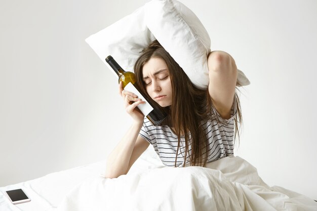 Sad young woman sitting on bed suffering from bad hangover after night party at club, having sleepy tired look, keeping eyes closed, holding bottle of wine and pillow, trying to cover ears from noise