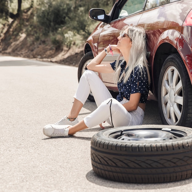 道路に壊れた車の近くに座って悲しい若い女性