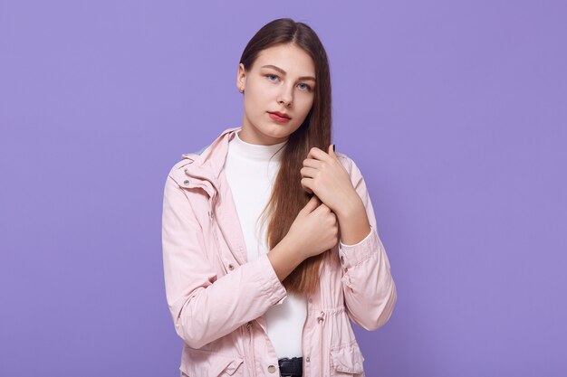 直接見ながら髪に触れる悲しい若い女性は、薄ピンクのジャケットと白いタートルネックを着て、薄紫色の壁の上に孤立して立って、動揺した表情をしています。