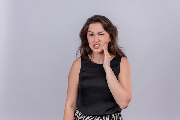 Sad young woman wearing black undershirt put her hand on aching tooth on white wall