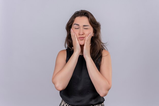 Sad young woman wearing black undershirt put her hands on cheeks on white wall