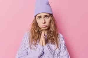 Free photo sad young woman with imploring look keeps palms pressed together begs for something needs your help and support wears purple loose jumper and hat isolated over pink background i am so sorry