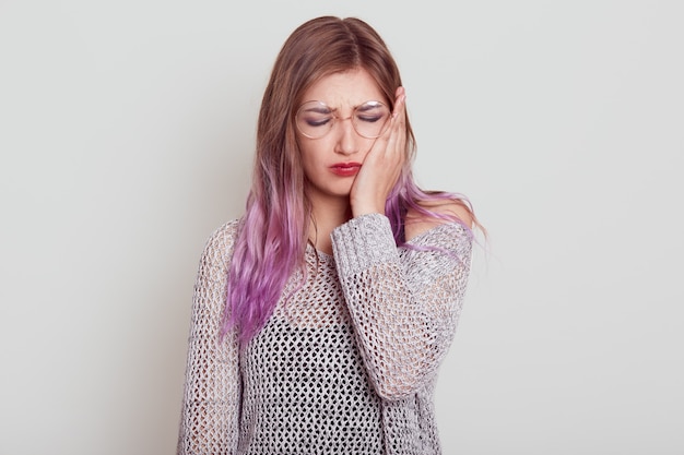 Sad young woman with lilac hair suffering from terrible toothache, touching her cheek with palm, keeps eyes closed, frowning face, isolated over grey background.