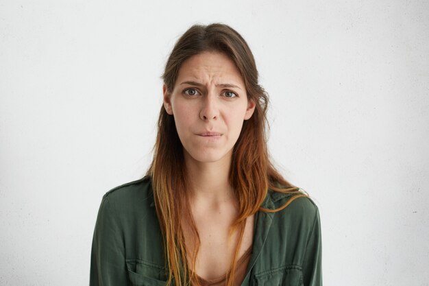 Foto gratuita triste giovane donna con lunghi capelli scuri, occhi affascinanti e pelle pura che curva le labbra guardando con disappunto avendo alcuni problemi al lavoro guardando sconvolto