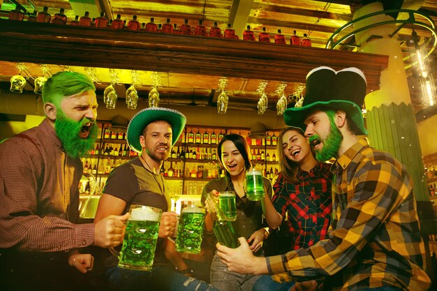 Saint Patrick's Day Party. Happy friends are celebrating and drinking green beer. Young men and women wearing green hats. Pub Interior.