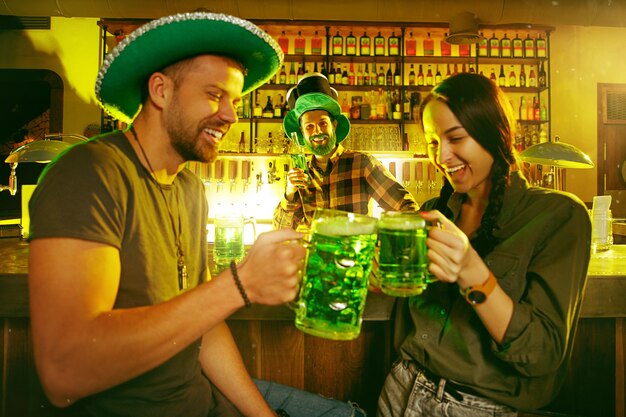 聖パトリックの日パーティー。幸せな友達が祝ってグリーンビールを飲んでいます。緑の帽子をかぶった若い男性と女性。パブのインテリア。