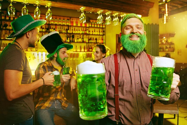 성 패트릭의 날 파티. 행복한 친구들은 축하하고 녹색 맥주를 마시고 있습니다. 녹색 모자를 쓰고 젊은 남성과 여성. 펍 인테리어.