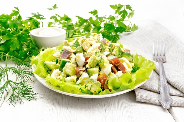 Salad from salmon, cucumber, eggs and avocado with mayonnaise on lettuce leaves in a plate, kitchen towel, dill, parsley and fork on a wooden board background