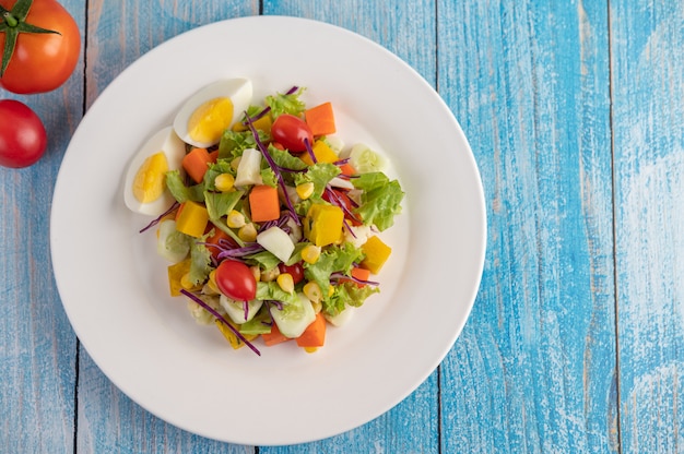 The salad is on a white plate, with a sandwich and tomatoes on the blue wooden floor.