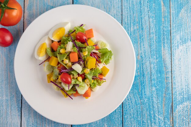 The salad is on a white plate, with a sandwich and tomatoes on the blue wooden floor.