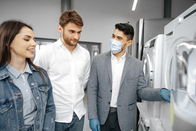 Salesman in hypermarket wearing medical mask demonstrates his clients a new washing machine