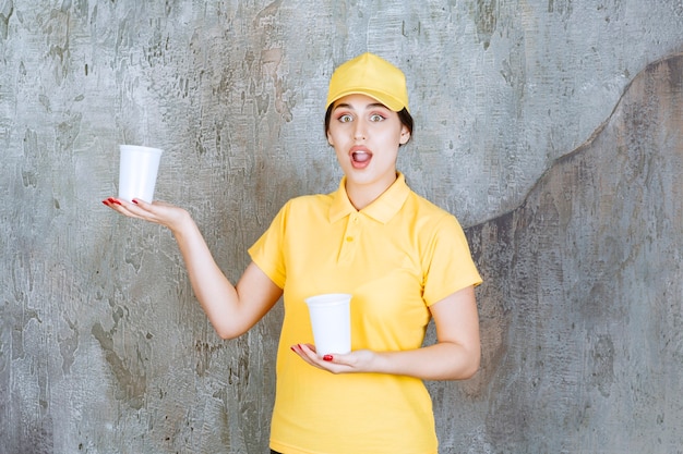 A saleswoman in yellow uniform holding two plastic cups of drink and giving one to the other person