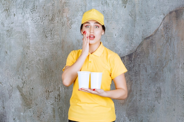 Free photo a saleswoman in yellow uniform holding two plastic cups of drink and looks stressed and terrified