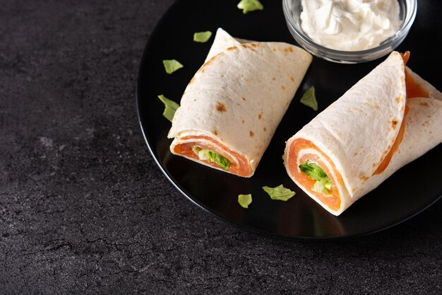 Salmon wrap sandwich roll with cheese and vegetables on black stone background