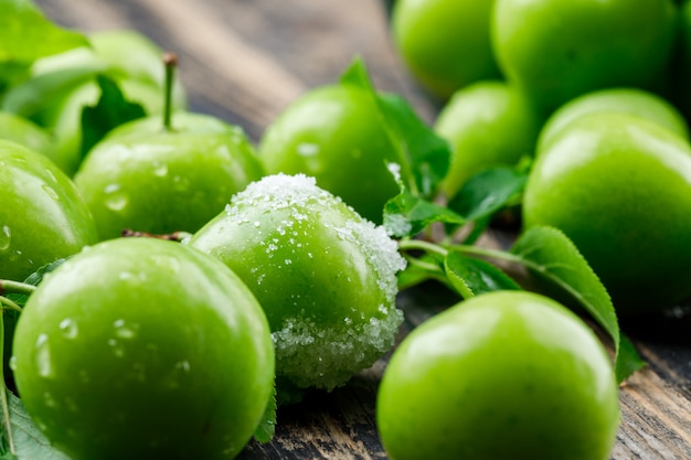 Salty green plums with leaves on wooden wall, close-up.