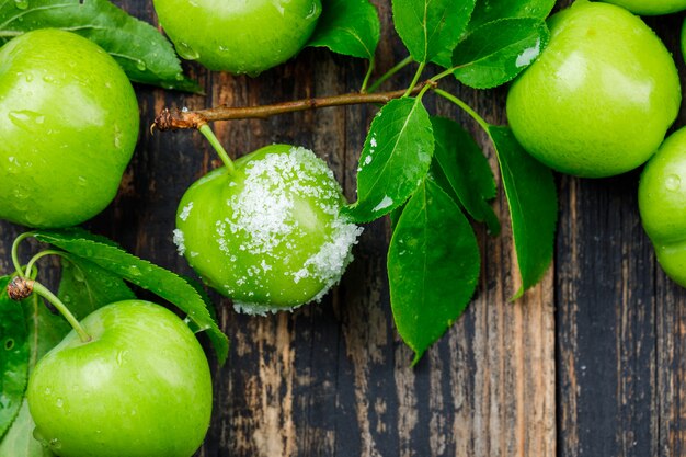 Salty green plums with leaves on wooden wall, flat lay.