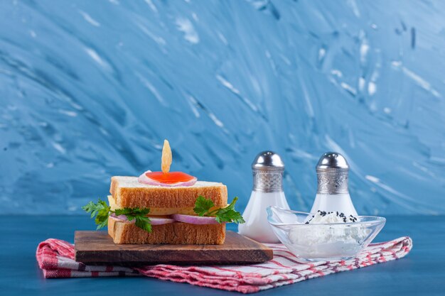 Sandwich on a board next to salt and a bowl of cheese on tea towel, on the blue table.