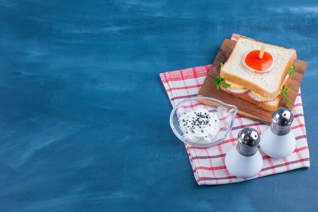 Sandwich on board next to salt and a bowl of cheese on tea towel on blue. 