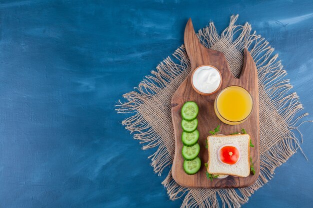 Sandwich, a glass of juice , bowl of cheese and sliced cucumber on a cutting board , on the blue background.