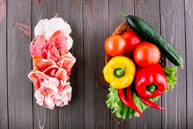 Sandwich made of slices of smoked meat and olives lies before basket with fresh vegetables