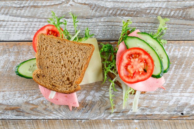 Sandwich with bread, cheese, tomato, cucumber, sausage, greens flat lay on a wooden table