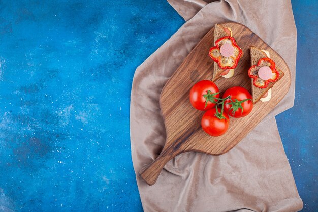 Sandwich with sausage and whole tomatoes on cutting board on pieces of fabric on blue. 