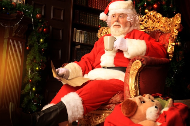 산타클로스는 집 벽난로 근처의 편안한 의자에서 휴식을 취하고 있습니다.