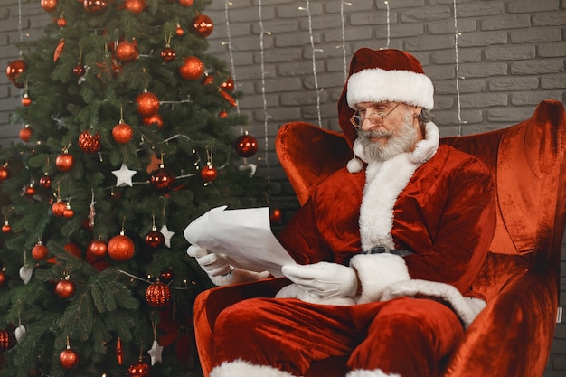 Santa Claus having a rest by the Christmass tree. Home decoration. Santa with letter from children.