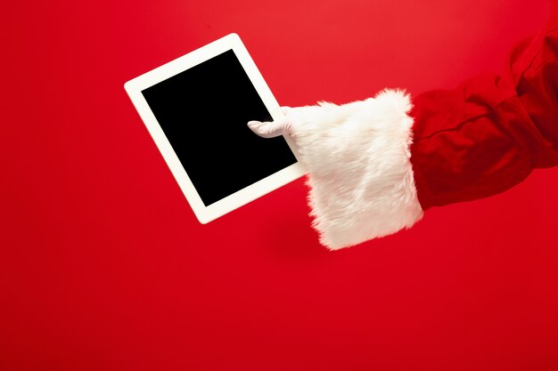 サンタクロースが赤いスタジオの背景にクリスマスの準備ができているラップトップを保持しています。季節、休日、お祝い、ギフトのコンセプト
