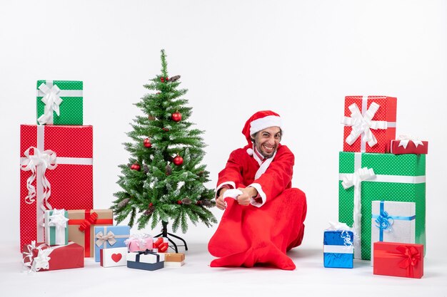 Santa claus sitting in the ground and holding christmas sock near gifts and decorated tree