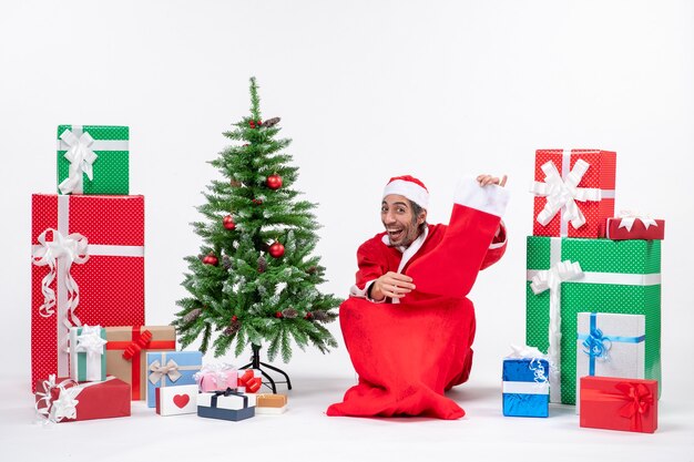 地面に座って、贈り物や飾られたクリスマスツリーの近くにクリスマスの靴下を見せているサンタクロース