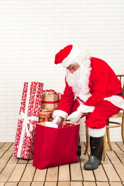 Санта-Клаус сидит и кладет подарки в бумажный пакет