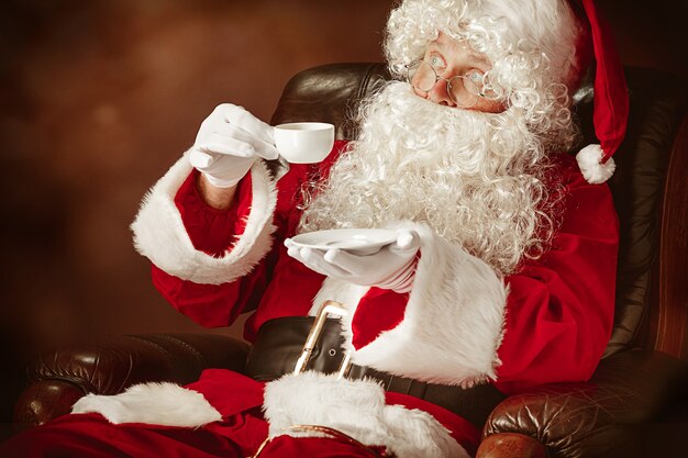 고급스러운 흰 수염, 산타의 모자 및 커피 한잔과 함께 의자에 앉아있는 빨간 의상을 입은 산타 클로스