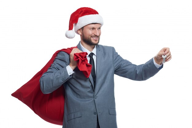 Santa clause in gray suite, red cap holding full bag over shoulder, smiling, looking away, giving key.