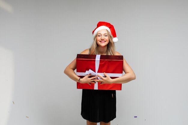 Santa girl in red hat with large festive gift smiling posing on gray background with copy space for xmas new year ad