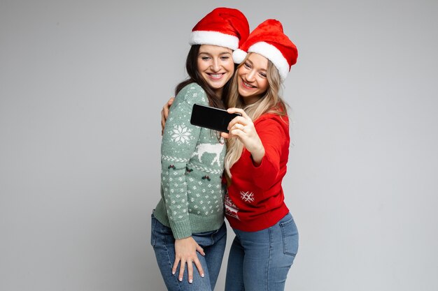 Santa girls friends in red hats posing with phone for festive selfie on gray background with copy space