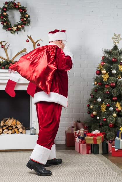 Santa with big sack of gifts behind back going to Christmas tree