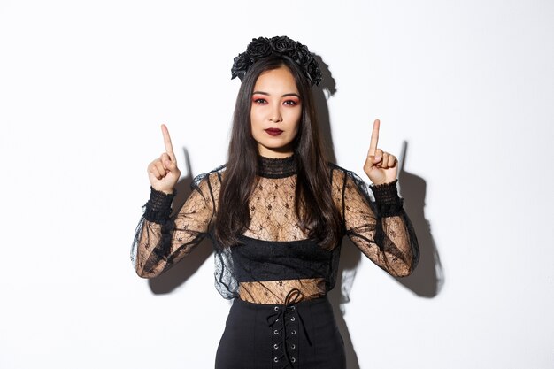 Sassy beautiful asian woman in black gothic dress, wearing witch costume for halloween and pointing fingers up