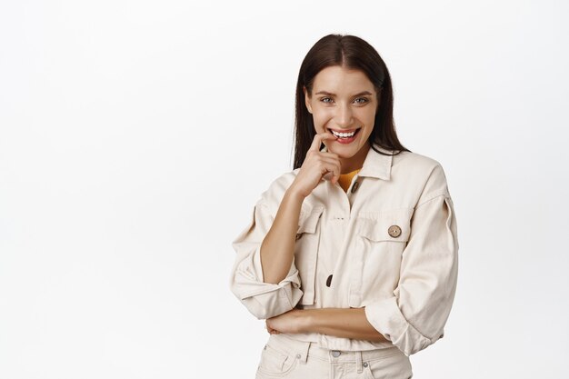 Sassy brunette woman, smiling flirty, biting finger and smiling cunning, has something in mind, thinking and glancing coquettish, standing  on white.