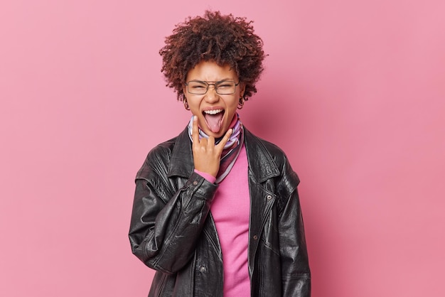 Sassy brunette young woman makes heavy metal gesture sticks out tongue enjoys favorite music on party wears spectacles black leather jacket poses against pink background Body language concept