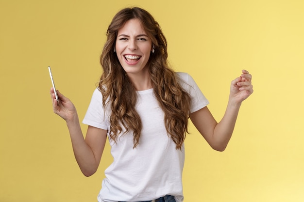 Sassy carefree joyful attractive woman having fun partying listen cool new song favorite artist dancing raise hands up squinting cheeky look camera lip sync hold smartphone yellow background