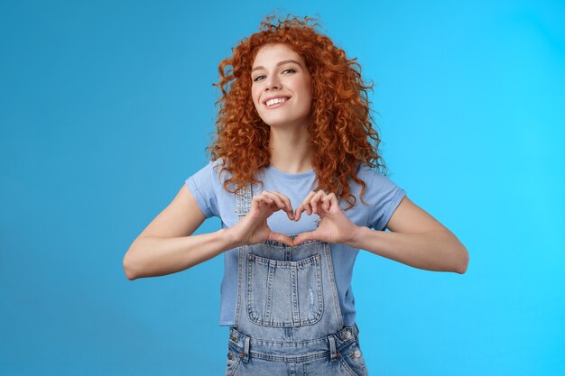 Sassy confident good-looking modern redhead curly woman raise head proud love tell girlfriend romantic heartwarming feelings present own heart smiling broadly standing blue background.