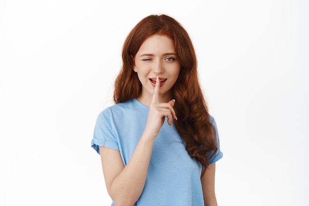 Sassy ginger girl yawning, hinting on secret, hiding something, make shush shh gesture tell to stay quiet, standing in t-shirt against white background