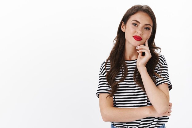 빨간 립스틱, 줄무늬 티셔츠, 흥미로운 아이디어를 듣고 턱을 만지고 기쁘게 웃는 현대 여성