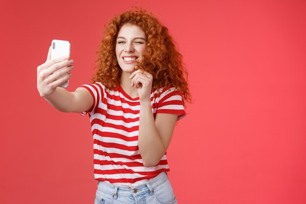 Sassy good-looking stylish charismatic redhead female curly hairstyle winking cheeky expression making flirty kinky faces hold smartphone taking selfie record video message play funny facial filters.