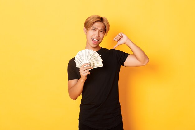 生意気なハンサムなアジアの男が現金で指を指し、満足そうに見えます。黄色の壁に立っている韓国人男性がお金を借りた
