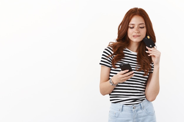 Нахальная современная милая молодая рыжая девушка прокручивает интернет-магазин, держит смартфон, кусает губу от соблазна, размышляет, что купить, держит кредитную карту, задумчиво смотрит на экран мобильного телефона, белая стена