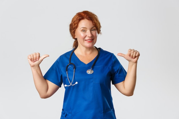 Sassy professional middle-aged doctor, female medical worker in scrubs pointing at herself and smiling, being skillful, 