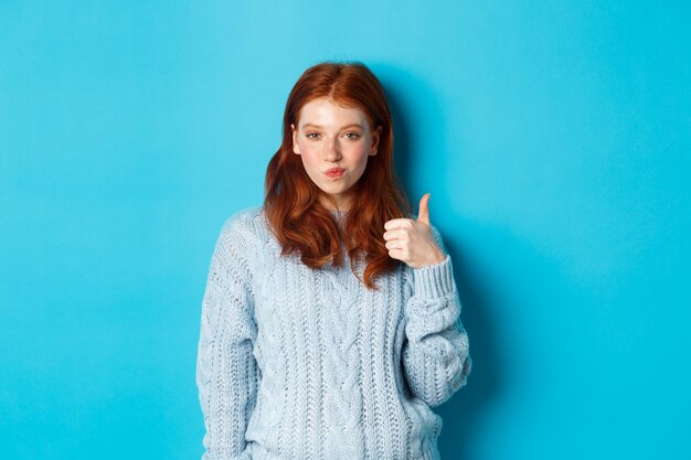 Sassy redhead girl in sweater, looking pleased and showing thumb up, like and agree, standing over blue background.